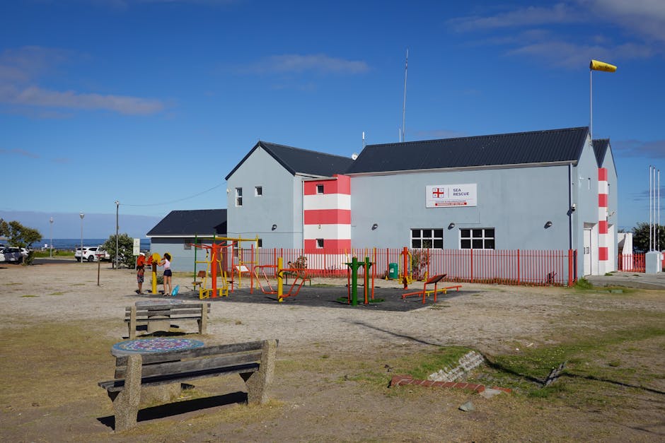 A grey and red Sea Rescue building with a black roof, situated in an open area with a gravel surface. In front of the building, there is an outdoor exercise area with colorful equipment, including yel