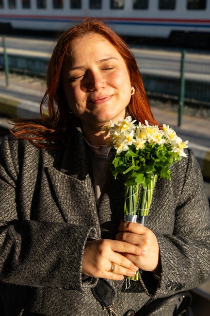 A woman with red hair and a gentle smile is holding a vibrant bouquet of fresh flowers, including yellow daisies and green foliage, wrapped in transparent glass. She is wearing a dark grey coat and st