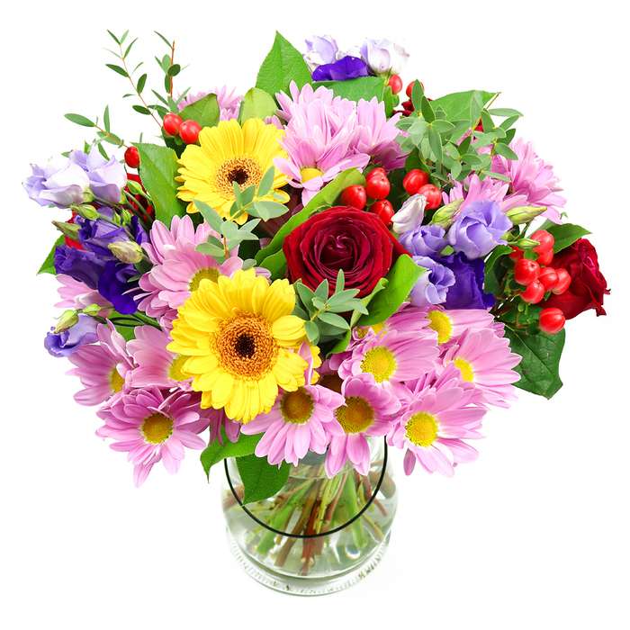 In this photograph, a vibrant mixed-colour bouquet fills a clear glass vase, its rounded shape and varied textures displayed against a clean white background for maximum clarity. Dominating the centre are lush deep red roses, their velvety petals layered in tight spirals that convey depth, emotion, and a hint of old-fashioned romance. Nestled beside them are two bright yellow gerbera daisies with dark brown-orange centres, their open faces lending the arrangement a sense of joy and spontaneity, like a celebratory gathering in a Gospel Oak café after a walk on Parliament Hill. Encircling these focal blooms, generous sprays of soft pink daisies or chrysanthemums form a cushion of pastel petals; each tiny yellow centre echoes the warmth of the gerberas, helping to tie the colours together in a harmonious, cheerful palette. Interlaced among the pink flowers, slender stems of lisianthus in shades of purple and gentle lavender rise gracefully, their bell-shaped flowers and buds adding movement, height, and a cool, elegant note that balances the warmer hues. Scattered throughout, rounded clusters of bright red hypericum berries bring a glossy texture and a slightly festive, modern accent. The bouquet is supported by an abundance of green foliage: broad, darker leaves give structure and richness, while finer, pale green sprigs create lightness and a natural, garden-picked feel. Through the clear glass vase, neatly cut stems fan into the water, revealing the skill and precision of an experienced florist. The overall impression is of an uplifting, generous arrangement, perfect for birthdays, thank-you gestures, or a house-warming gift for someone settling near Lissenden Gardens, adding colour, fragrance, and a sense of thoughtful celebration to everyday life in Gospel Oak.